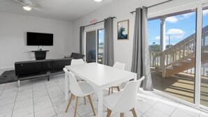 Un espace repas moderne avec une table et des chaises blanches, un canapé noir, un téléviseur à écran plat et une vue sur un escalier et un espace extérieur.