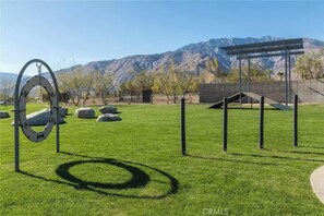 A grassy outdoor area with a metal structure, rocks, and a mountain backdrop. - Luxe Desert Escape on the Grove w/ 360° Mountain Views (Palm Springs)