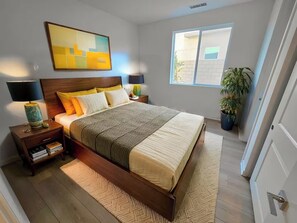 A modern bedroom with a large bed, wooden headboard, bedside tables with lamps, a floor lamp, and a window with a view of the outside. - Luxe Desert Escape on the Grove w/ 360° Mountain Views (Palm Springs)
