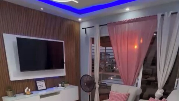A modern living room with a flat-screen TV, a white sofa with pink cushions, a ceiling fan, a large window with curtains, and a staircase visible through the window. - Elegant & Comfortable Apartment with Private Balcony (Santo Domingo Este)