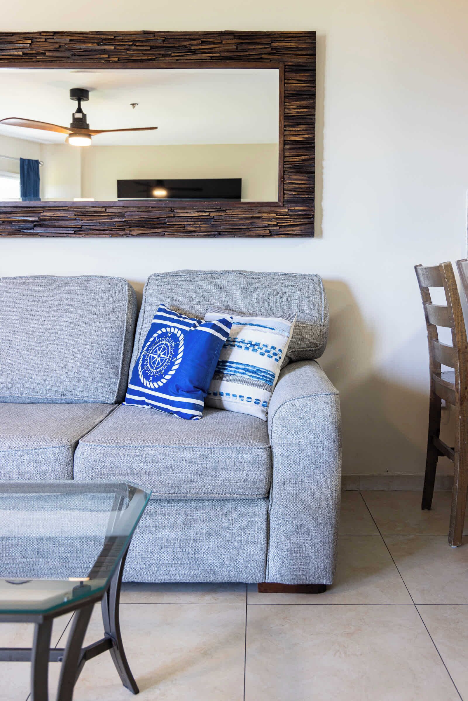 Un sofá gris con dos almohadas azules y blancas estampadas, una mesa con superficie de vidrio y una silla de madera.