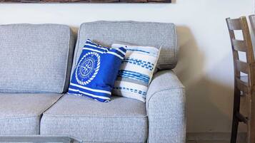 Un sofá gris con dos almohadas azules y blancas estampadas, una mesa con superficie de vidrio y una silla de madera.