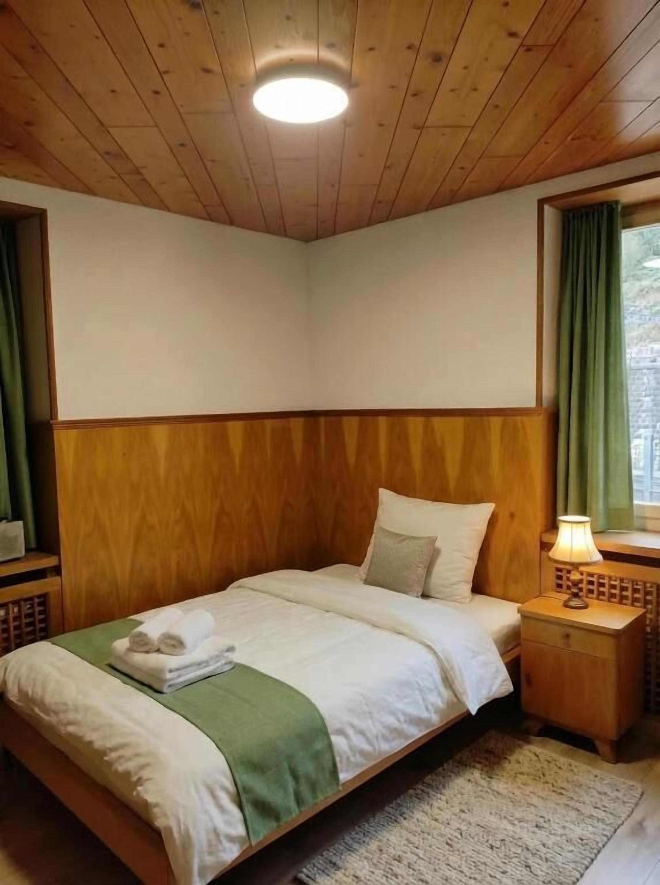 A bedroom with a wooden ceiling, a bed with white bedding, a green throw, a nightstand with a lamp, and a window with curtains.