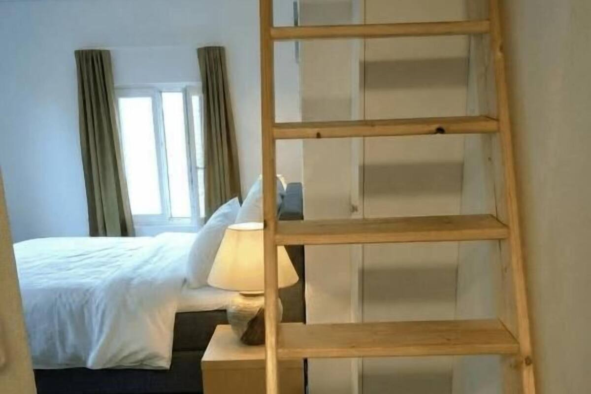 A bedroom with a wooden ladder, a bed, a nightstand, and a window with curtains.