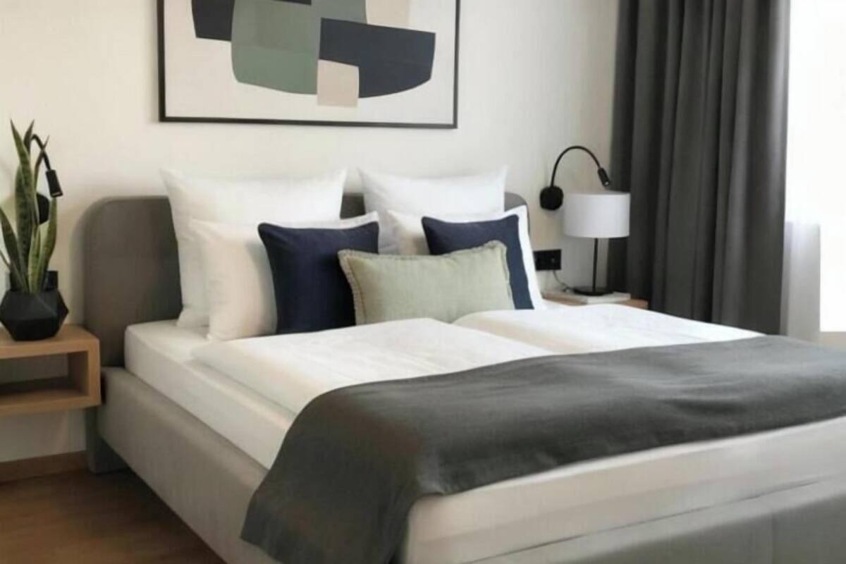 A neatly made bed with white and dark blue pillows, a grey blanket, and a modern abstract painting on the wall.