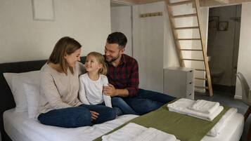 Eine dreiköpfige Familie auf einem Bett mit einer grünen Decke und weißen Handtüchern, einer Holzleiter und einer weißen Wand mit einem kleinen Bilderrahmen.