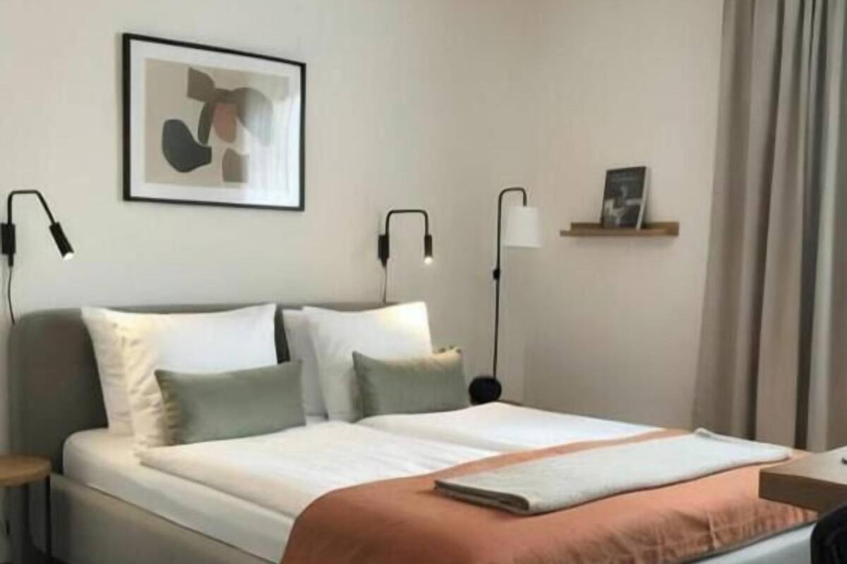 A neatly made bed with a white and orange blanket, two bedside lamps, and a framed abstract artwork on the wall.