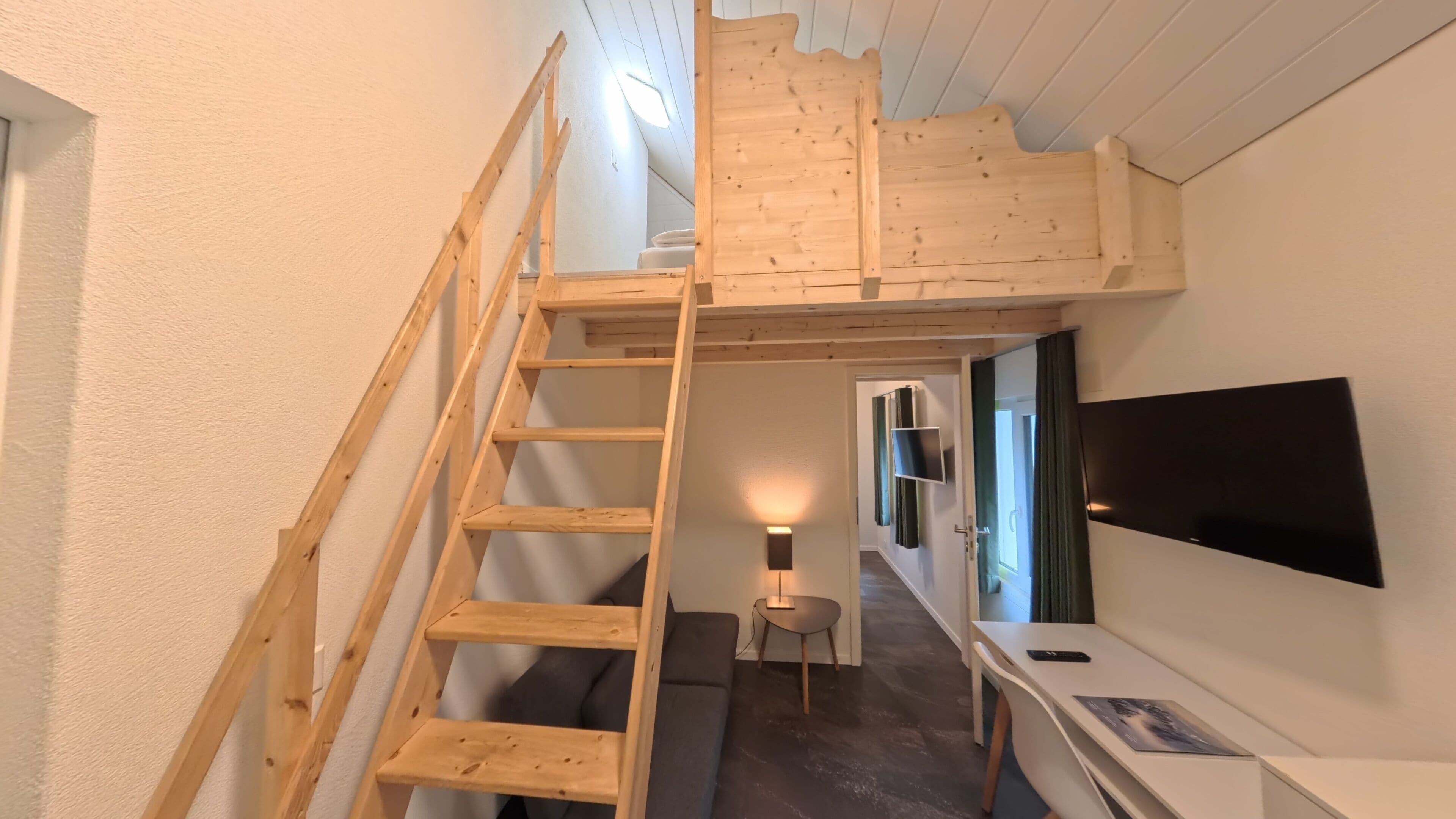 Ein kompakter Raum mit einer Holztreppe, die zu einer Schlafgalerie führt, einem Schreibtisch mit Stuhl, einem an der Wand befestigten Flachbildfernseher und einem kleinen Tisch mit Lampe.
