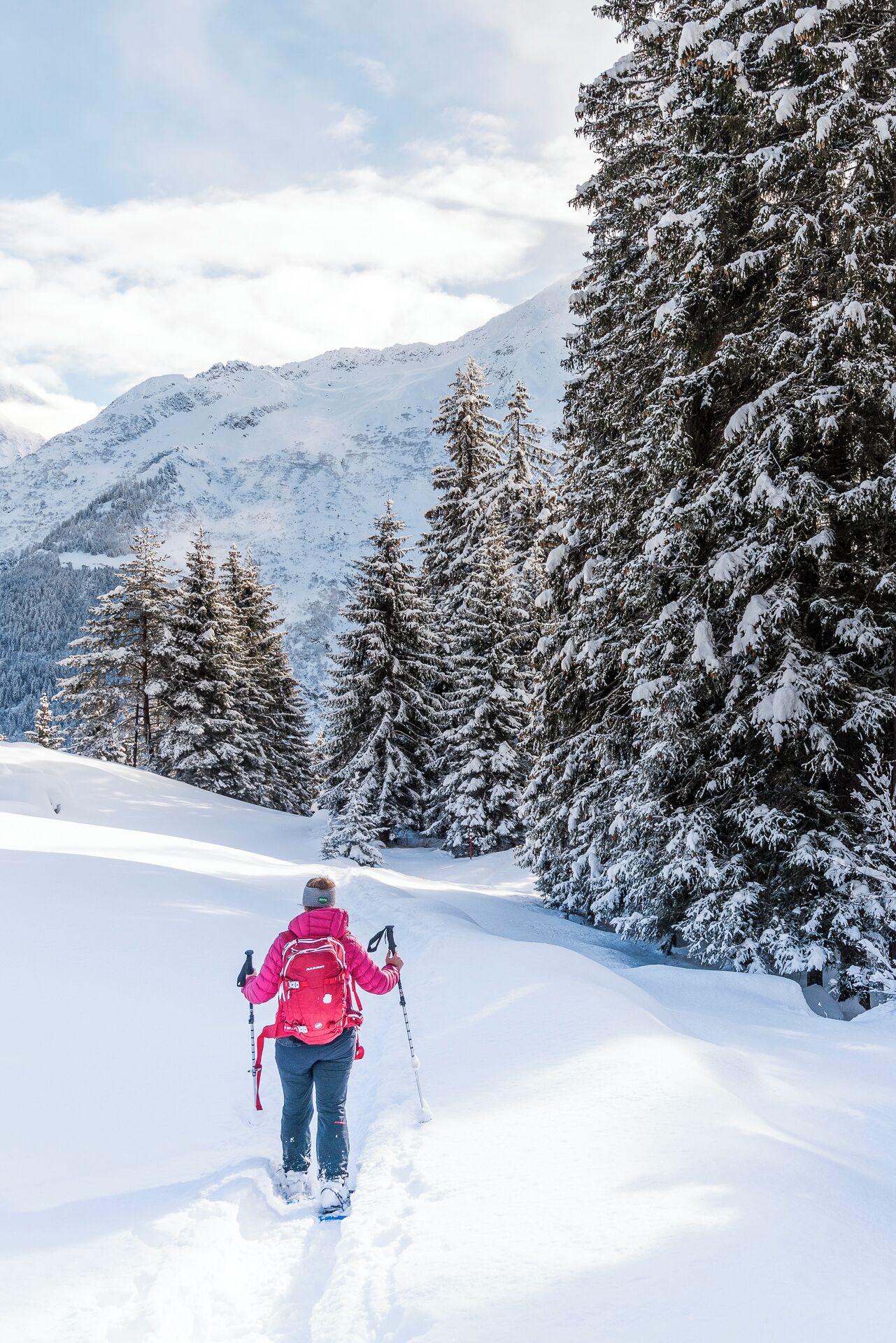 A snowy mountain landscape with a person in a pink jacket and red backpack walking away from the camera.