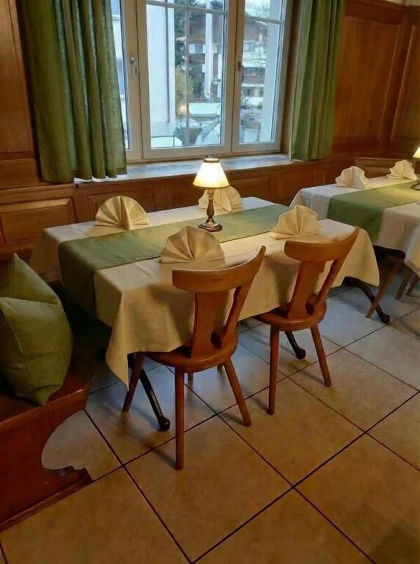 A dining area with tables set for two, a lamp on one table, and a view of buildings through large windows.