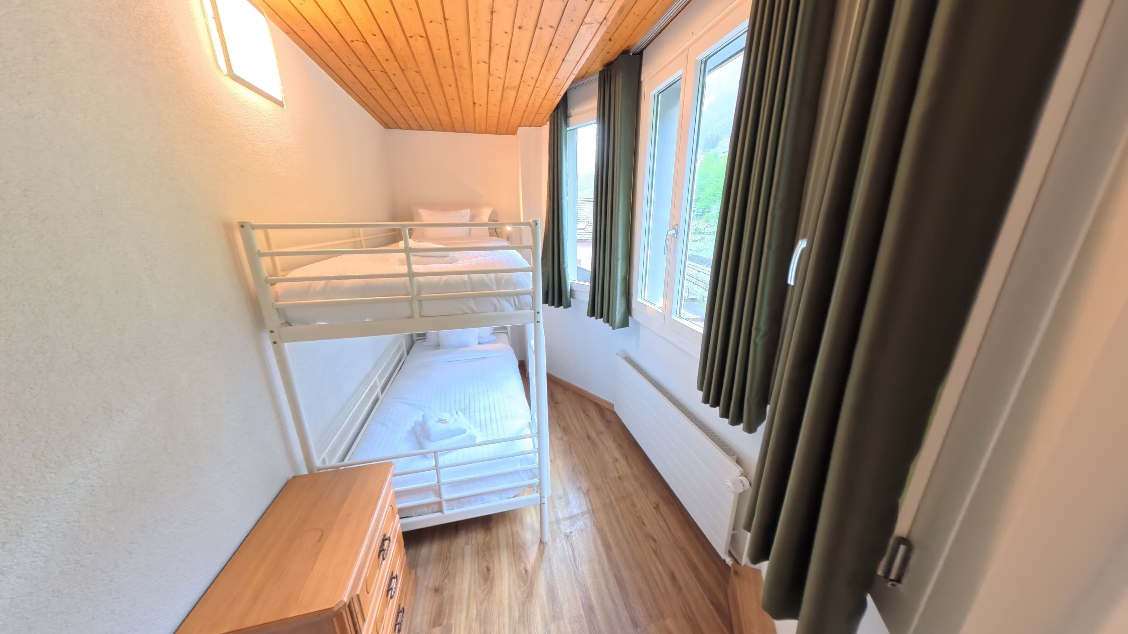 A bunk bed room with a wooden ceiling, a window with curtains, and a wooden bench.