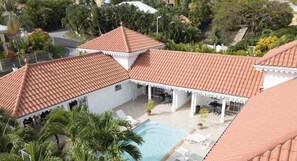 Zona de piscina en la azotea con techo de baldosas, rodeada de palmeras y un edificio blanco con balcón.