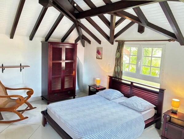 Un dormitorio con cama, mecedora de madera, armario y ventana con cortinas.