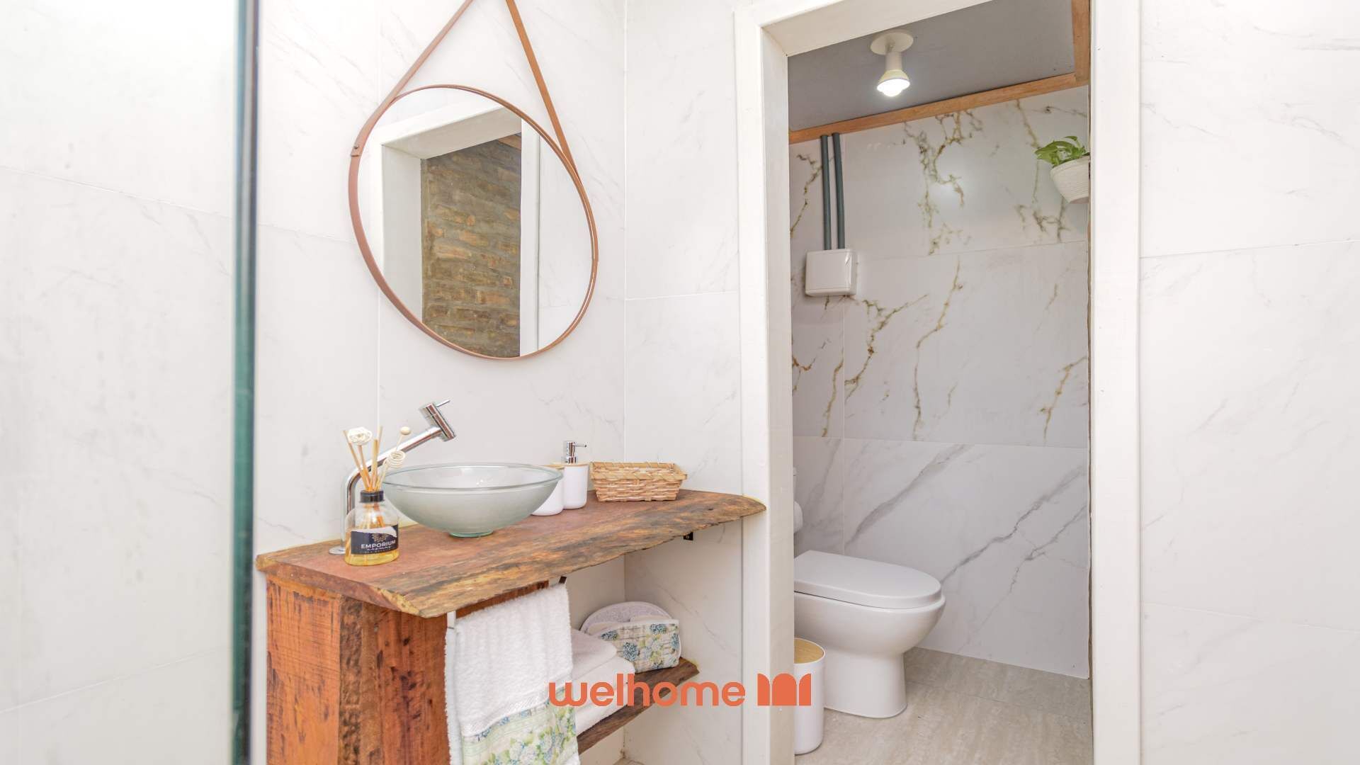 Ein Badezimmer mit Marmorwand, Waschtisch aus Holz, einem Waschbecken aus Glas, Toilette und einem hängenden Spiegel.