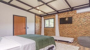 A bedroom with a brick wall, a flat-screen TV, a bed with a green blanket, a bench, and a window with curtains.