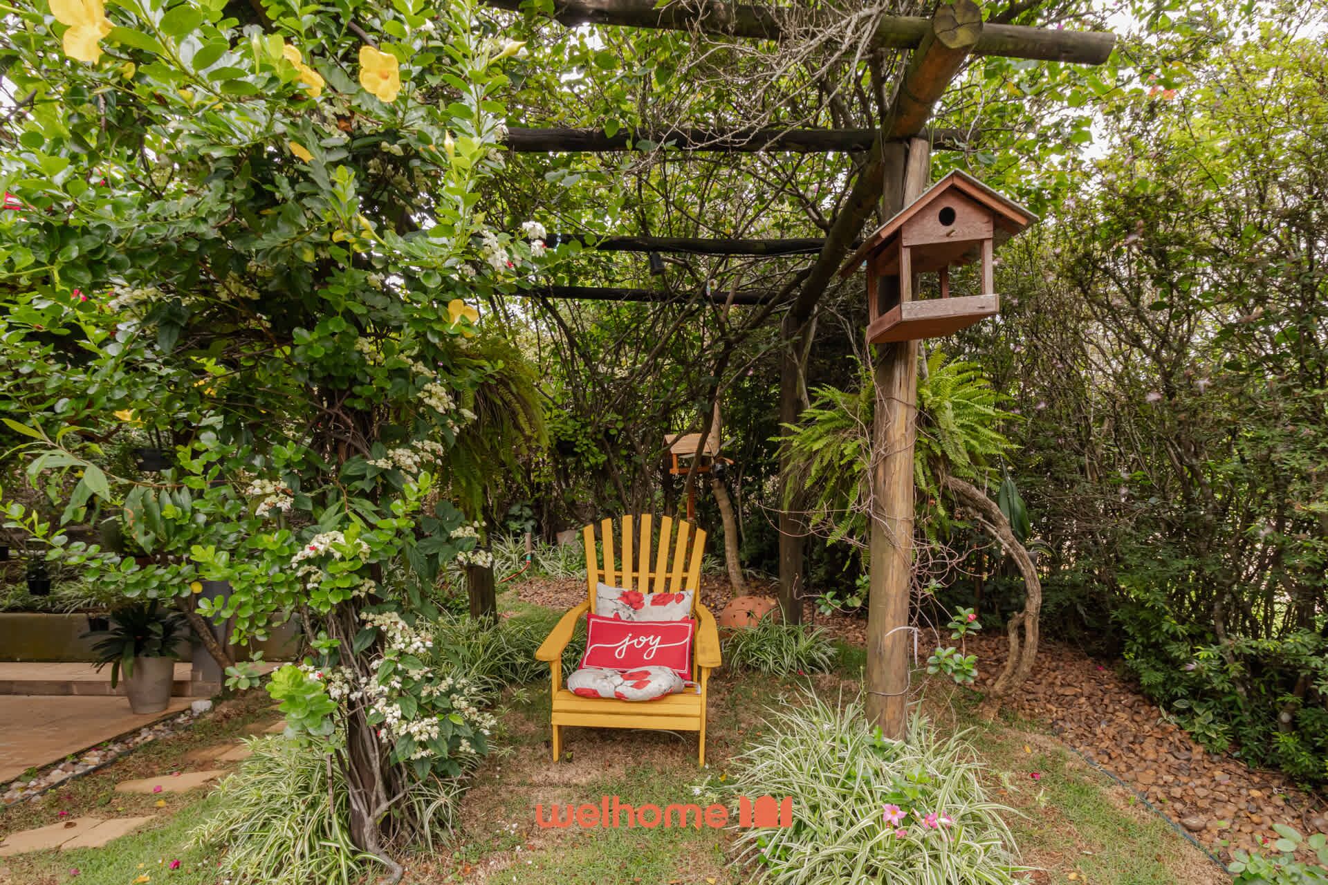 A wooden chair with a cushion and a pillow with the word "joy" on it, placed in a garden with a birdhouse hanging from a wooden structure.