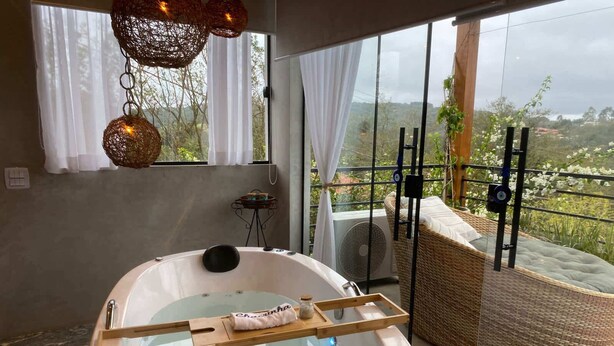 A modern bathroom with a freestanding bathtub, a wicker chair, and a view of the outdoors. - Sitio em Ibiuna com piscina próximo rota do vinho (ibiuna)