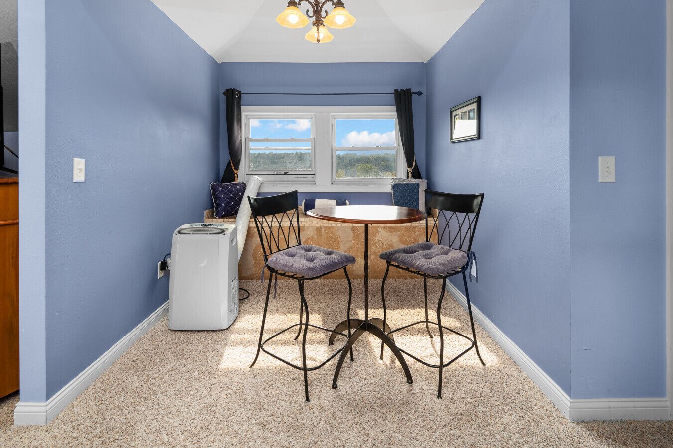 A dining area with a round table and four chairs, a window with curtains, and a ceiling light fixture.