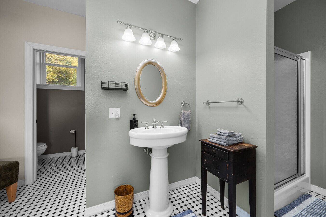 A bathroom with a white pedestal sink, a round mirror with a gold frame, and a towel rack.