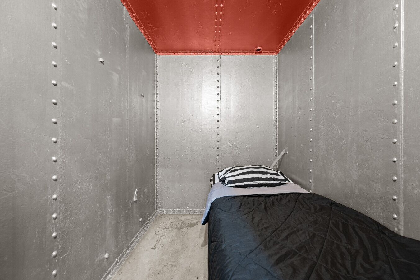 A small, narrow room with a single bed, a striped pillow, and metal walls with rivets.