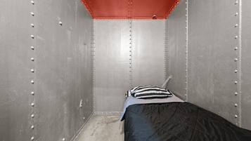 A small, narrow room with a single bed, a striped pillow, and metal walls with rivets.