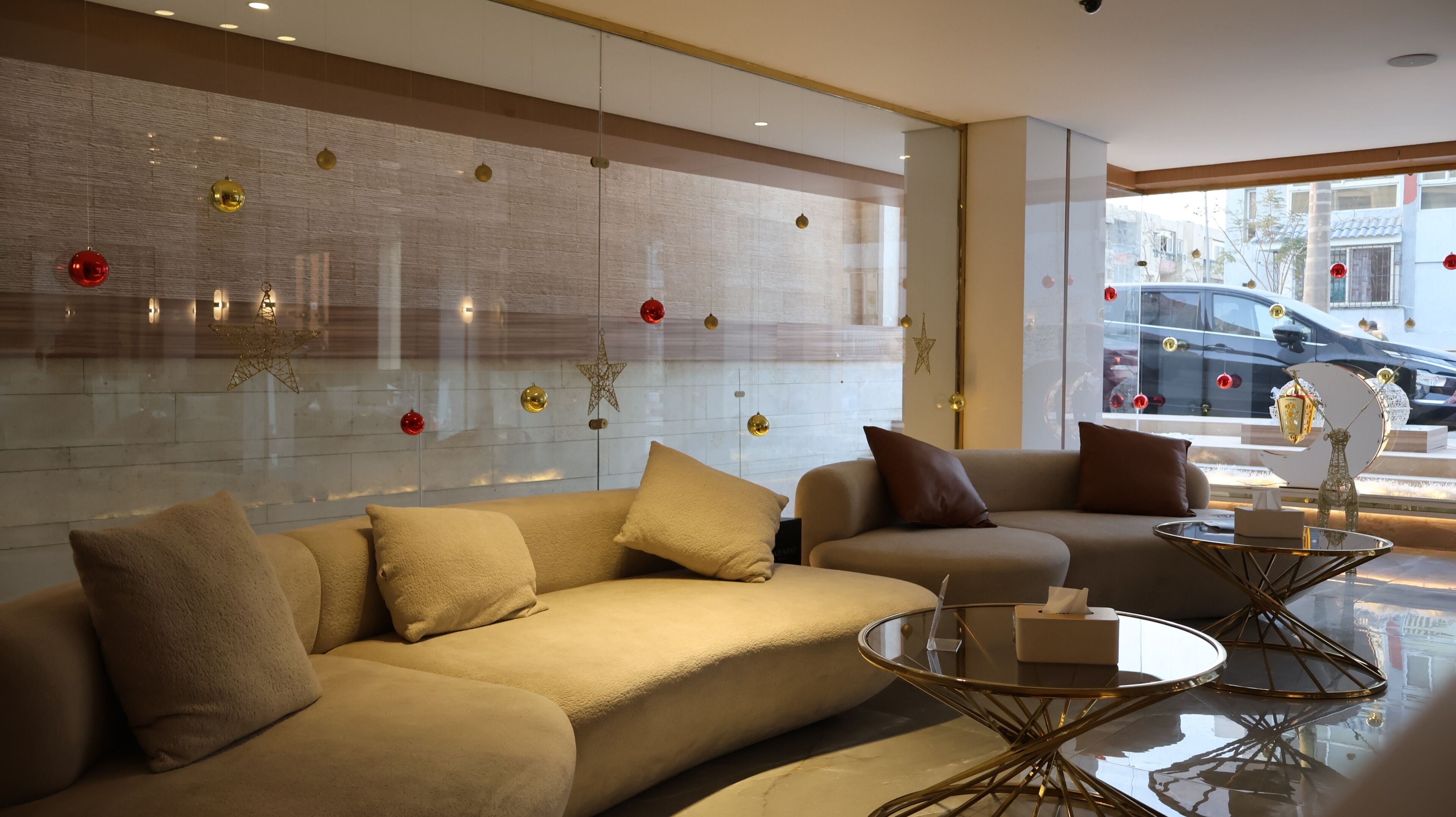 A modern living room with a beige sofa, a round glass coffee table, and a large window with a view of the street.
