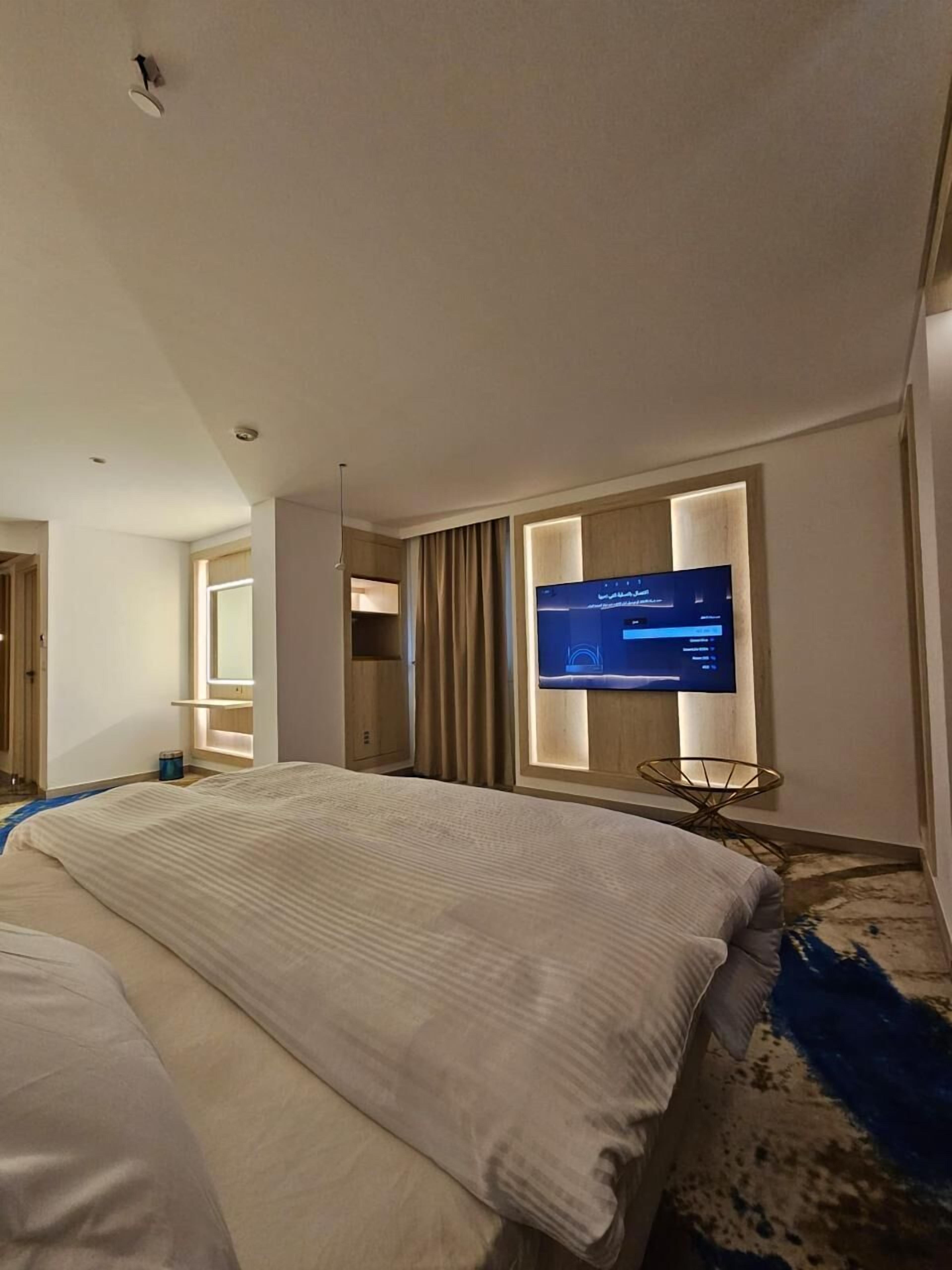 Una habitación de hotel con una cama grande, un televisor de pantalla plana en la pared, una mesita y una ventana con cortinas.