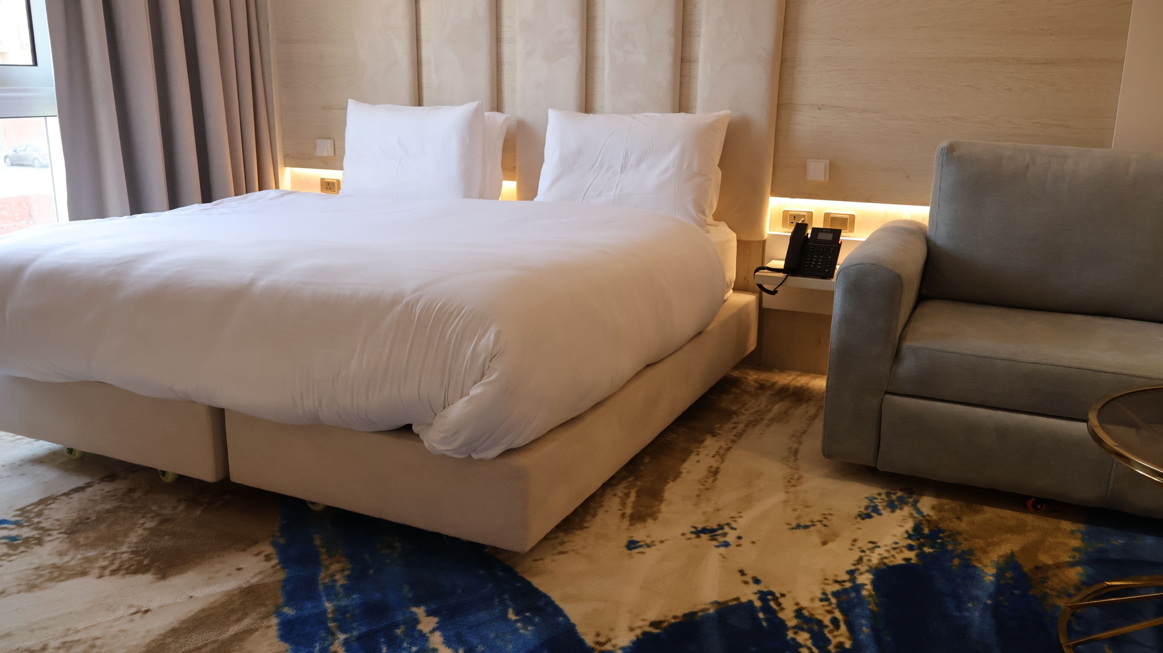 Quarto de hotel com uma cama grande, uma mesinha de cabeceira com telefone e uma poltrona cinza.