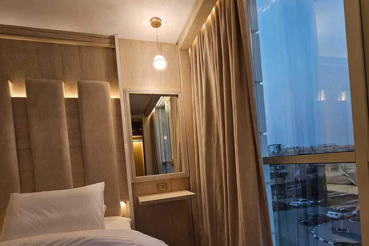 Habitación de hotel con una cama grande, una mesita de noche, un espejo y vistas a un aparcamiento a través de un ventanal amplio.