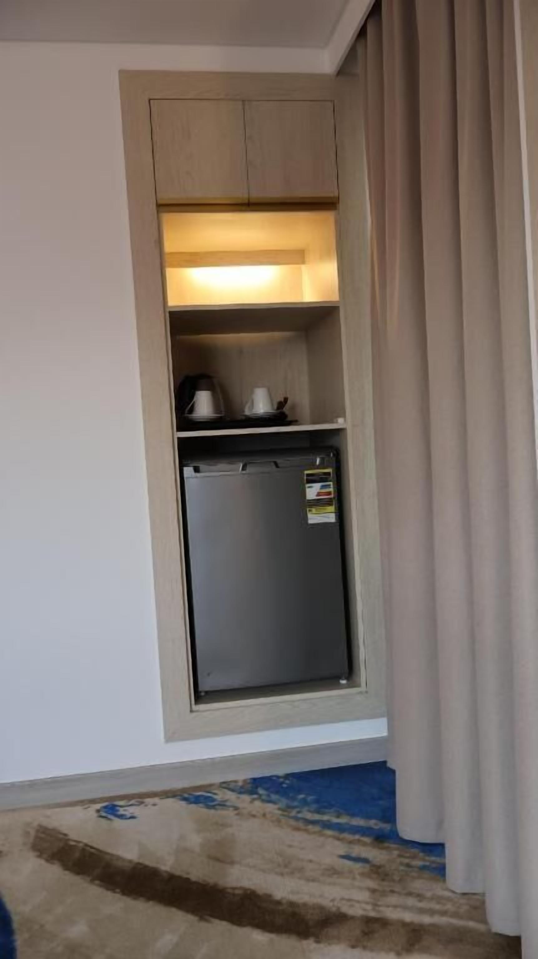 A compact room with a built-in refrigerator, a small shelf with cups, and a patterned rug on the floor.