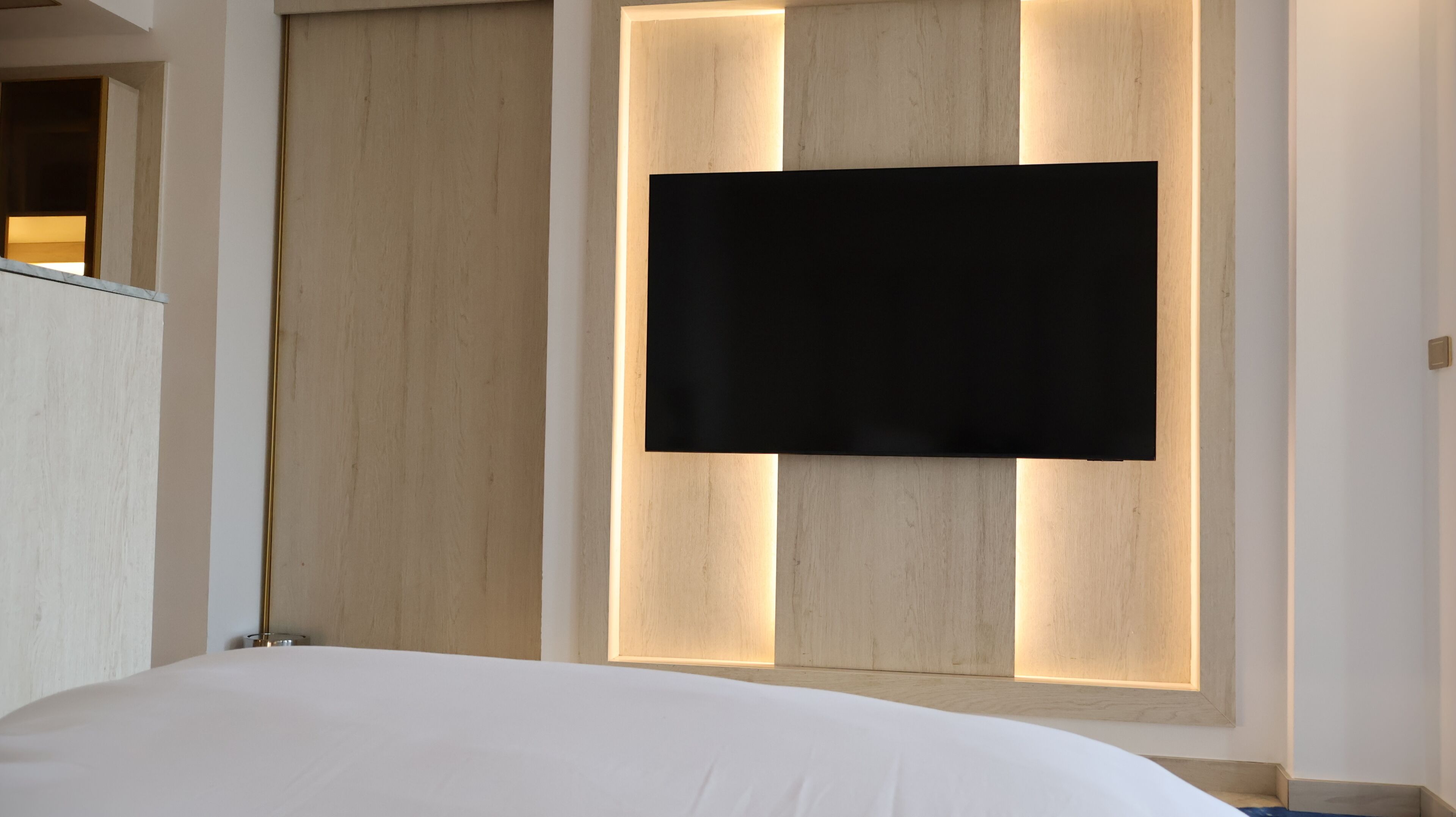 Una habitación de hotel moderna con una cama grande, un televisor de pantalla plana montado en una pared de paneles de madera y un armario de madera.