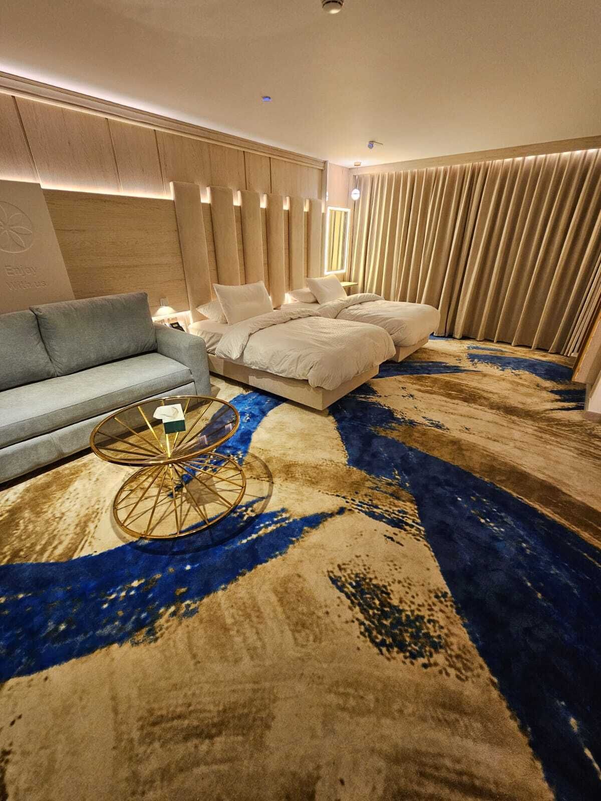 Una habitación de hotel moderna con sofá, cama con ropa de cama blanca, alfombra estampada y una mesa de centro original.