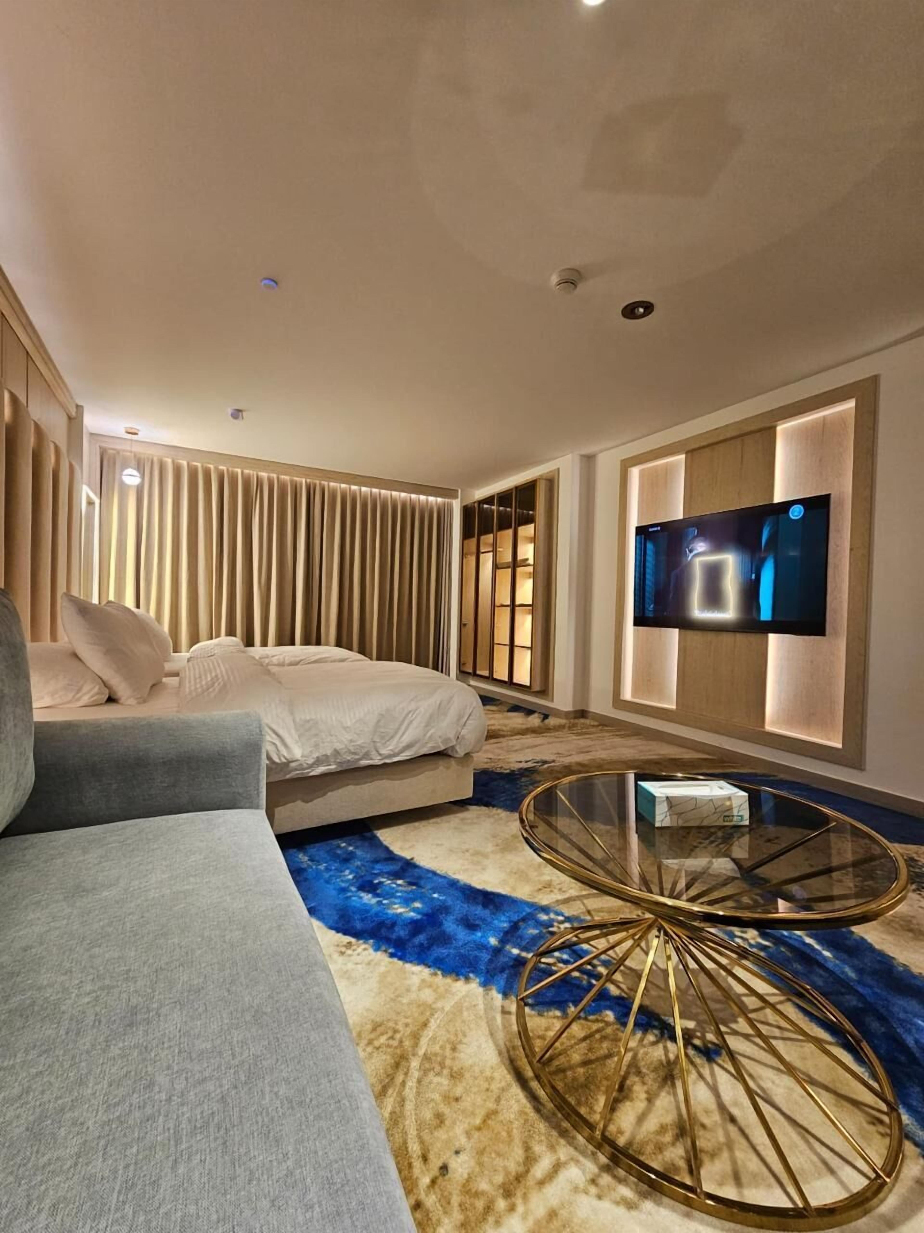 Quarto de hotel moderno com cama, sofá, televisão fixada na parede e uma mesa de centro de vidro singular.