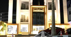 La façade d'un hôtel Sheraton de nuit, avec une enseigne lumineuse, de grandes fenêtres et une entrée bien visible.
