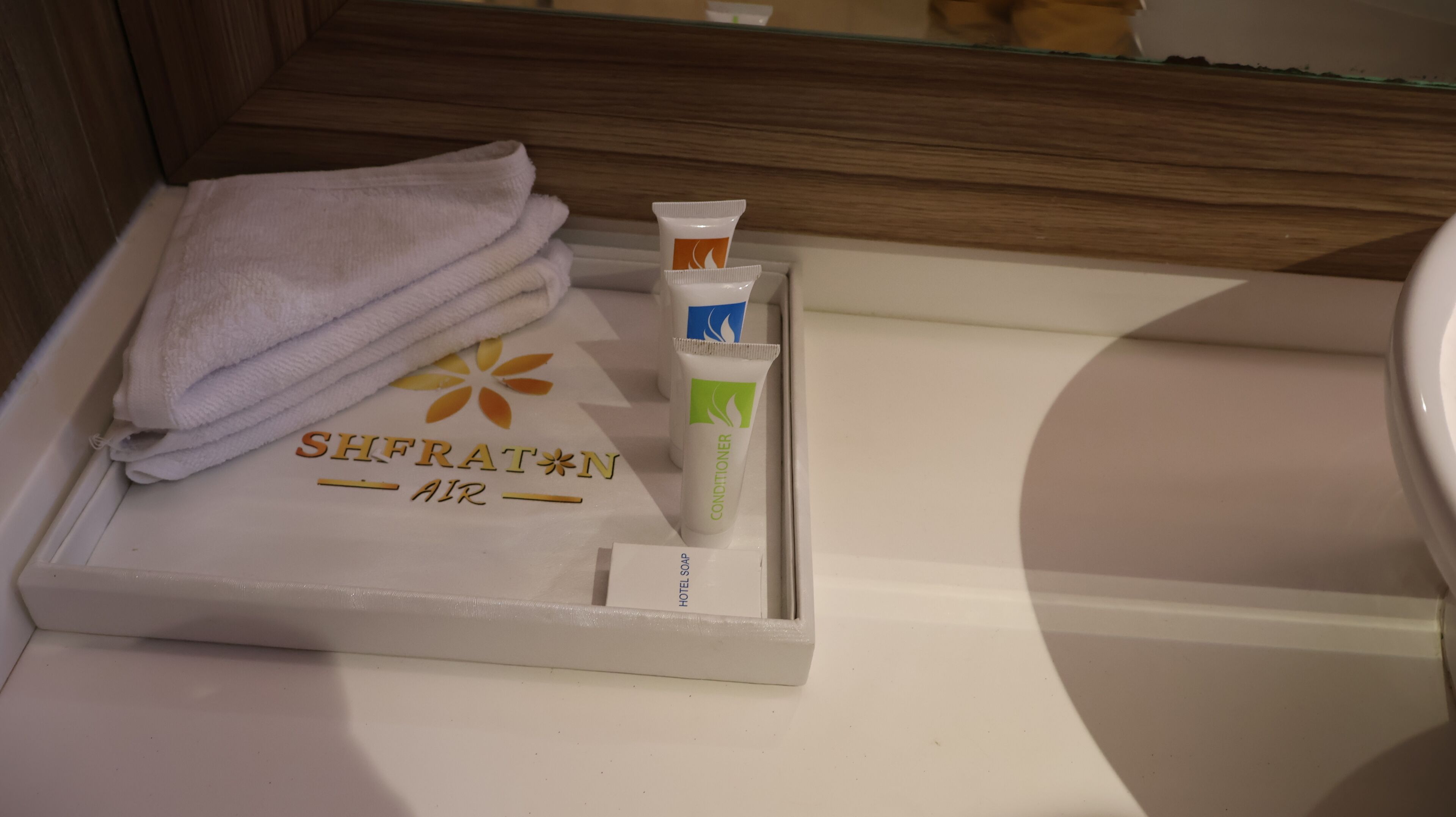 Un baño con una toalla, un tubo de acondicionador y una caja con la etiqueta 'SHFRATIN AIR' en un estante.
