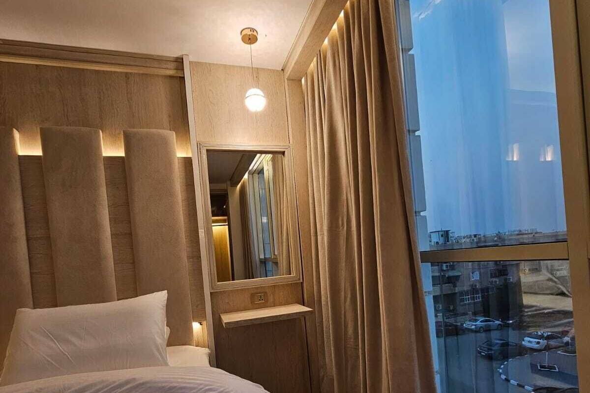 Habitación de hotel con una cama grande, una mesita de noche, un espejo y vistas a un aparcamiento a través de un ventanal amplio.