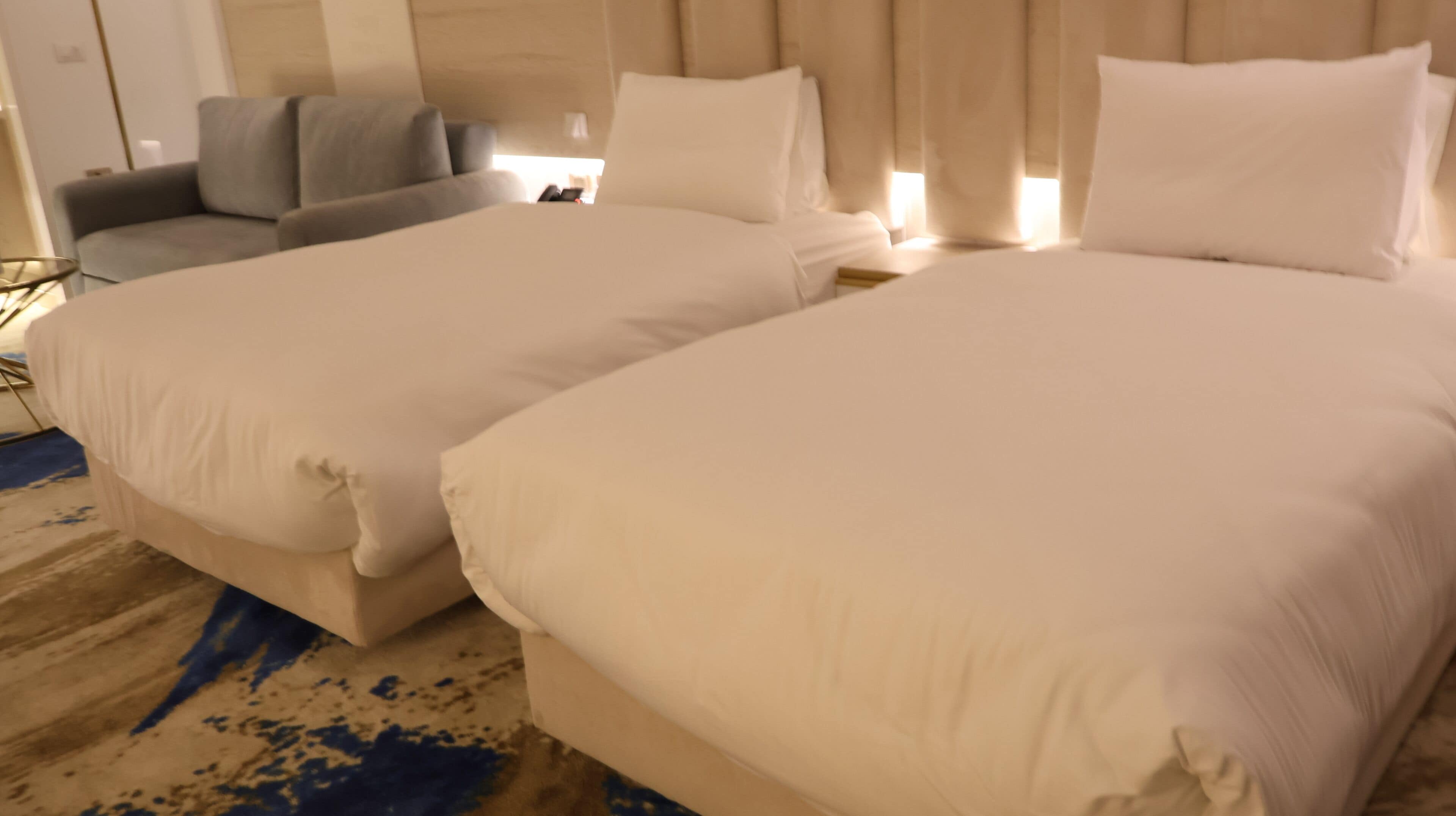 A hotel room with two beds, a grey armchair, and a large window with curtains.