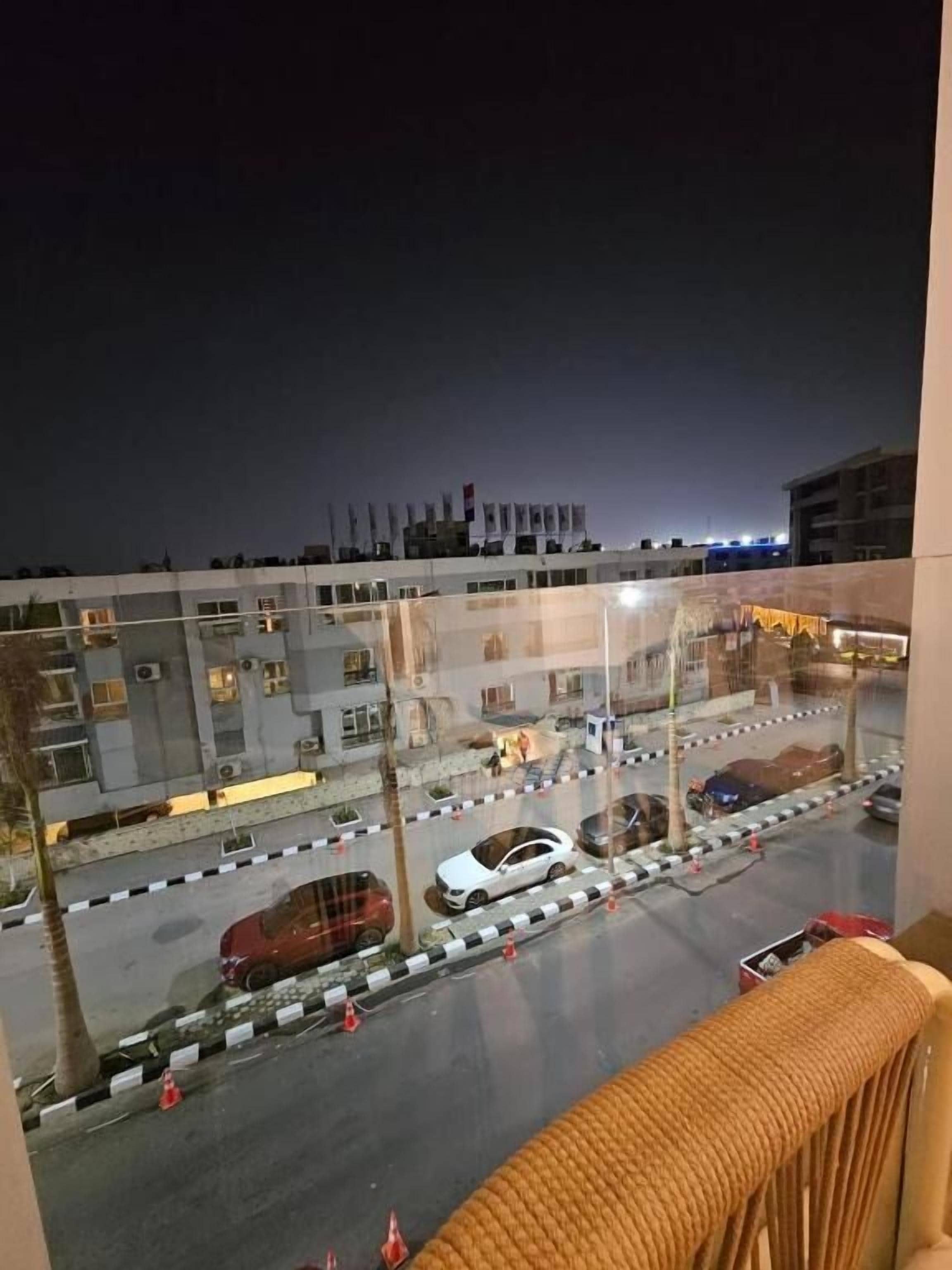 ’s Nachts uitzicht vanaf een balkon op een parkeerplaats met meerdere auto’s, een gebouw met verschillende verdiepingen en een verre stadshemel.