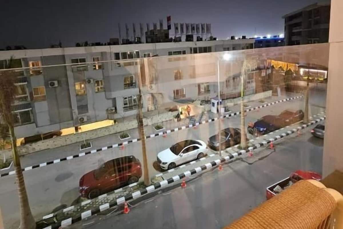 Vista nocturna desde un balcón sobre un estacionamiento con varios autos, un edificio de varios pisos y un horizonte urbano distante.
