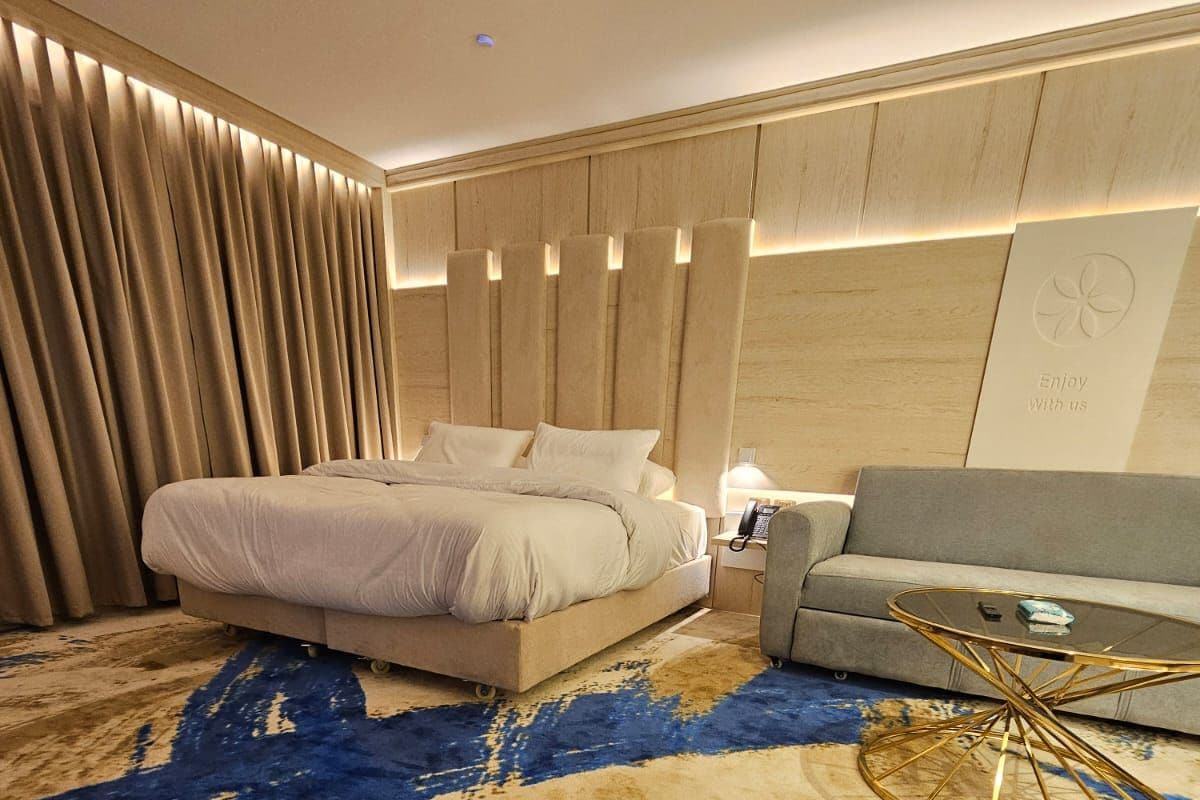 A modern hotel room with a large bed, a sofa, a glass coffee table, and a patterned carpet.