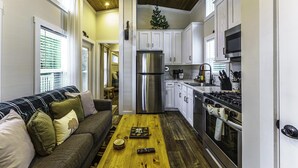 Une cuisine compacte équipée d’un réfrigérateur, d’une cuisinière et d’un four à micro-ondes. On y trouve une table en bois avec une télécommande et un objet décoratif. Un canapé avec des coussins est placé contre le mur, et un petit sapin de Noël est suspendu au plafond.