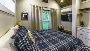 Une chambre à coucher comprenant un lit, une table de chevet, une fenêtre avec des rideaux, un téléviseur fixé au mur et une petite étagère avec quelques objets.