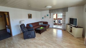 A living room with a sofa, a chair, a coffee table, a TV on a cabinet, and a sliding glass door leading to an outdoor area. - Eichweg vacation apartment (Bad Sooden-Allendorf)