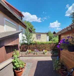 Een binnentuin met een geplaveid pad, plantenpotten, een houten hek en een gebouw met een raam en een deur.