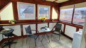 A covered patio area with a ceiling fan, two windows with views of a house, a small table with chairs, and a refrigerator. - Eichweg vacation apartment (Bad Sooden-Allendorf)
