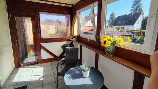 A room with a tiled floor, a black chair, a small round table with a potted plant, and a window with a view of houses and trees. - Eichweg vacation apartment (Bad Sooden-Allendorf)