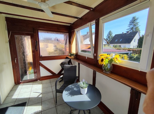 Una habitación con piso de baldosas, una silla negra, una pequeña mesa redonda con una planta en maceta y un ventanal con vistas a casas y árboles.