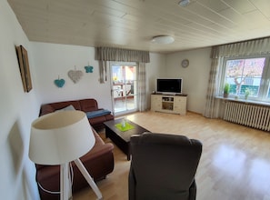 A living room with a sofa, armchair, coffee table, TV stand, and a window with curtains.