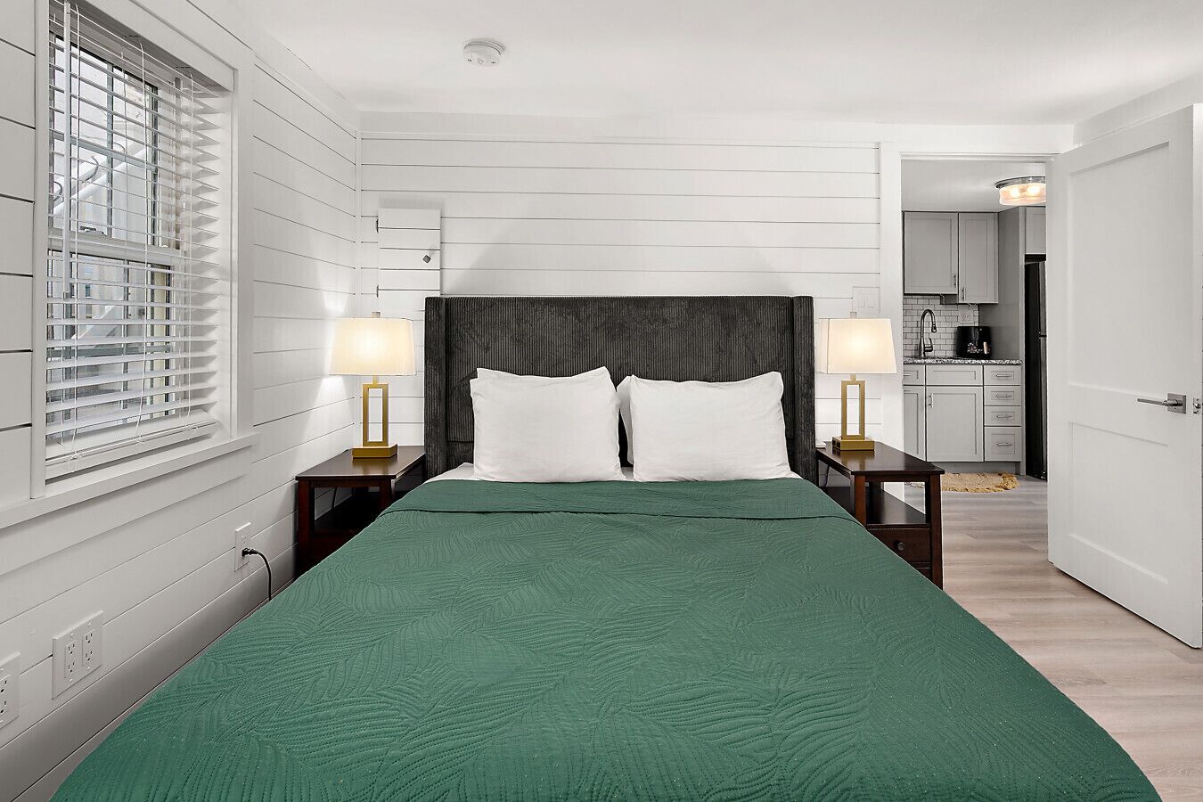 Quarto de hotel com uma cama, duas luminárias de cabeceira, uma janela com persianas, uma área de cozinha visível por uma porta aberta e uma porta branca.
