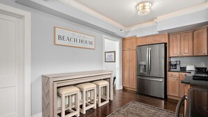 A kitchen with wooden cabinets, a stainless steel refrigerator, and a bar area with stools. - Surf 1 - Steps to the beach and boardwalk (Ocean City)